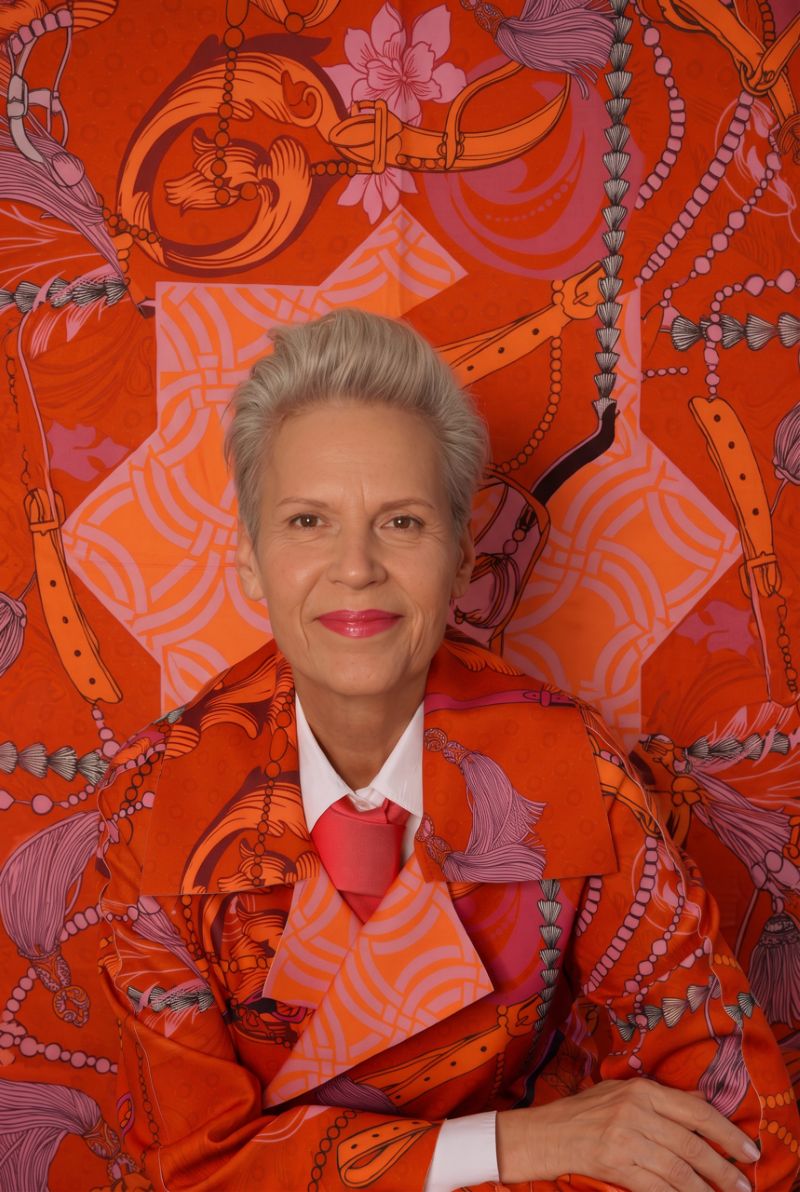 A person with short, gray hair is smiling and wearing a bright orange-and-pink patterned outfit that blends harmoniously with the matching background, creating a striking visual effect.