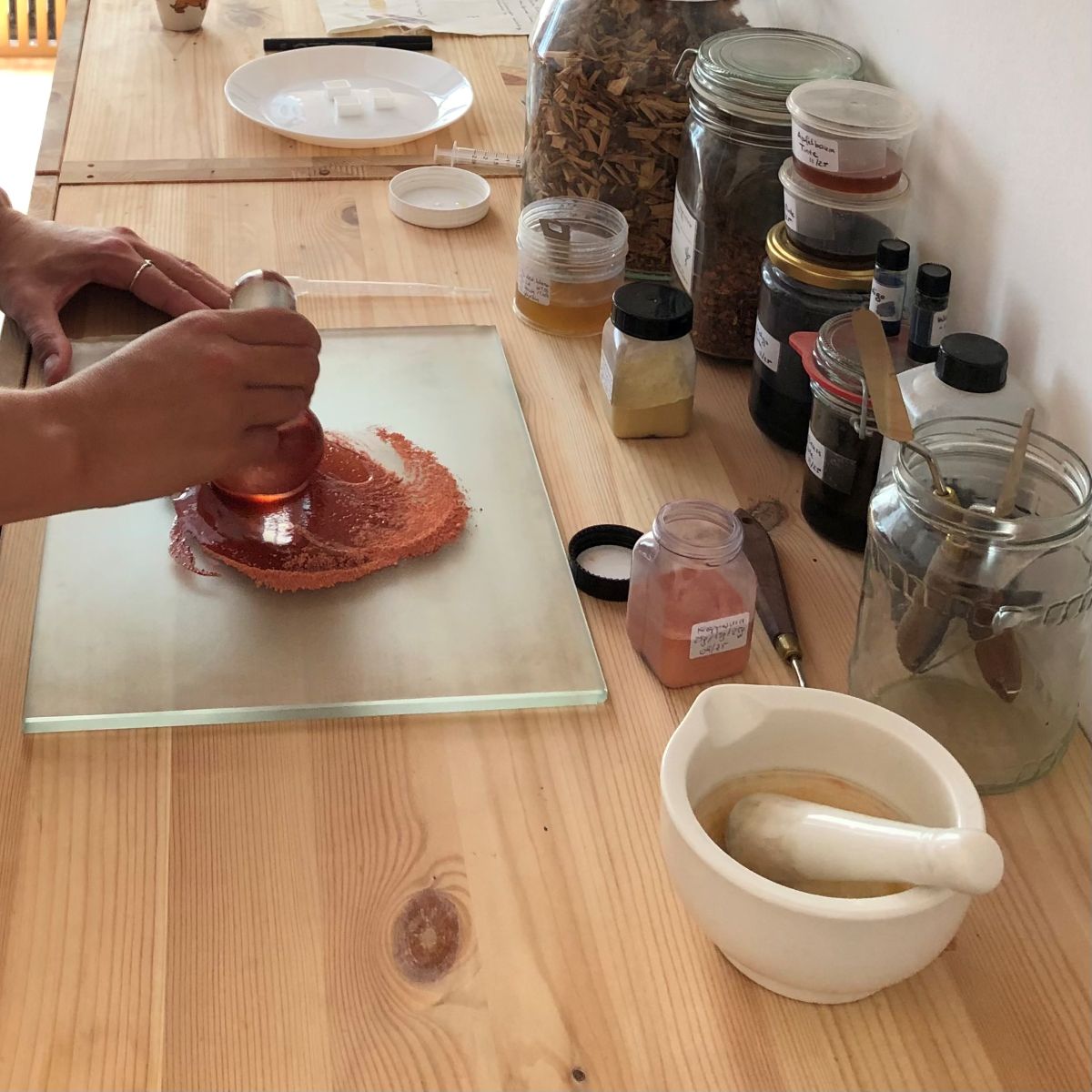 A reddish powder is ground with a pestle on a glass plate; on a wooden table sit spice jars, a mortar and pestle, and various containers that evoke a sense of community and the creation of creative worlds in the kitchen.