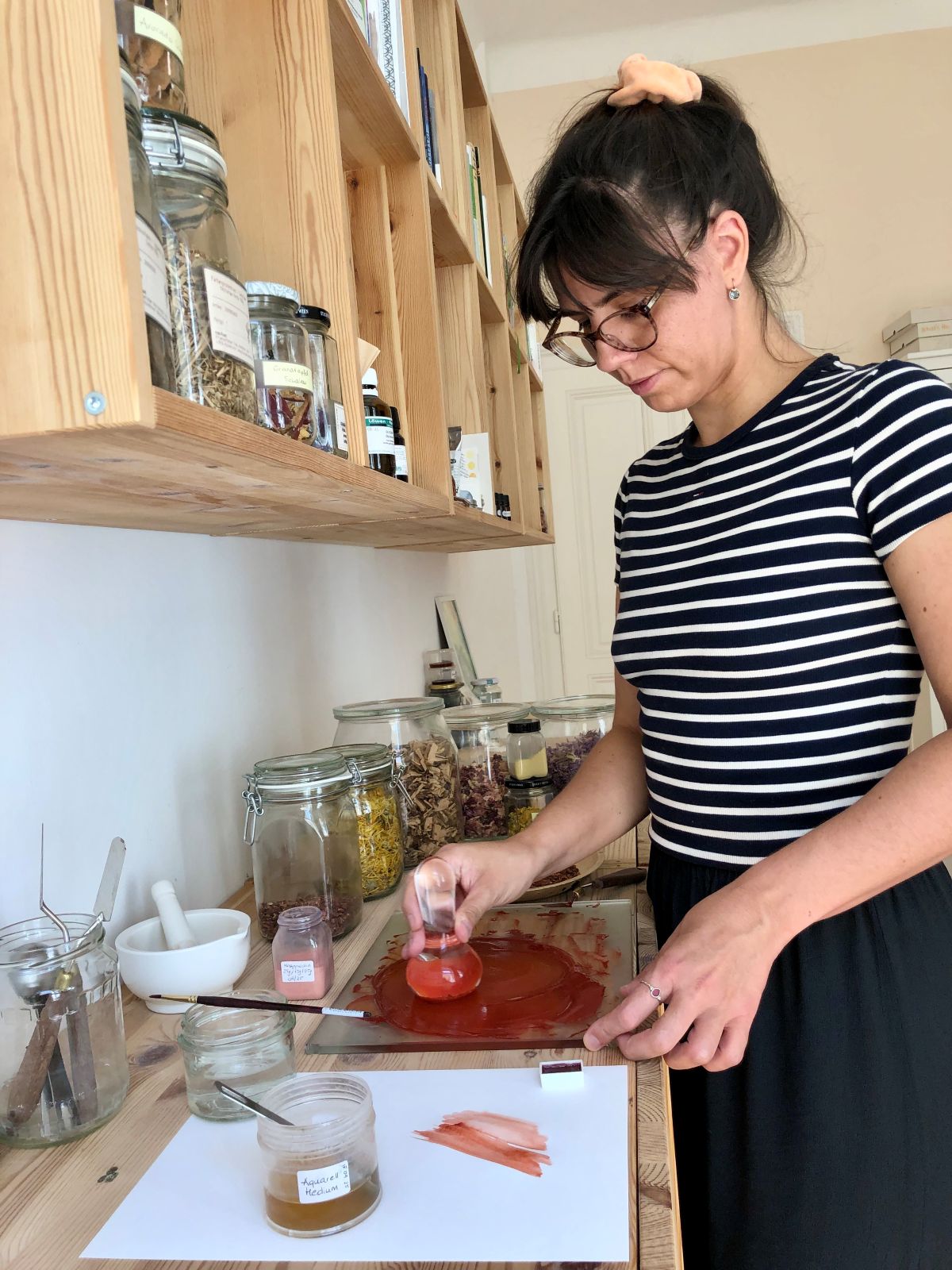 In a brightly lit room, a woman wearing glasses and a striped shirt mixes paint on a glass palette, surrounded by jars filled with various materials and ingredients, as she immerses herself in creative worlds and draws inspiration from the spirit of community.