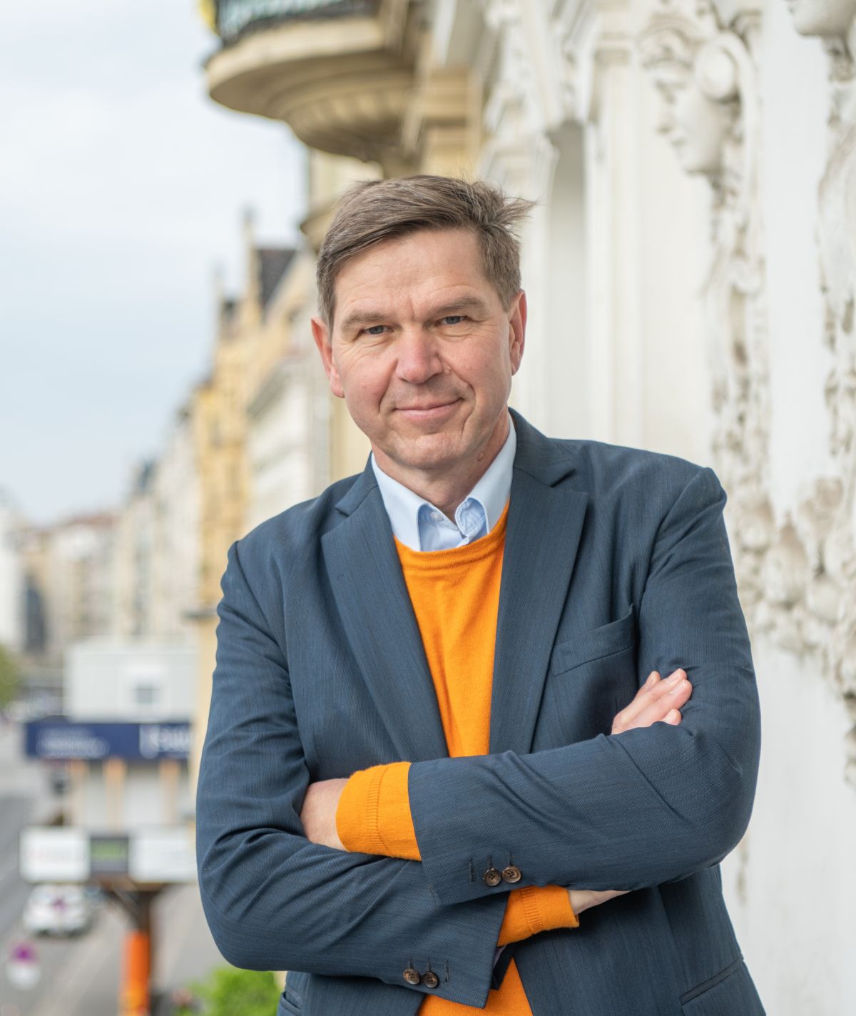 A man in a blue suit and an orange sweater stands smiling with his arms crossed on a balcony featuring ornate white architecture; a blurry city street can be seen in the background.