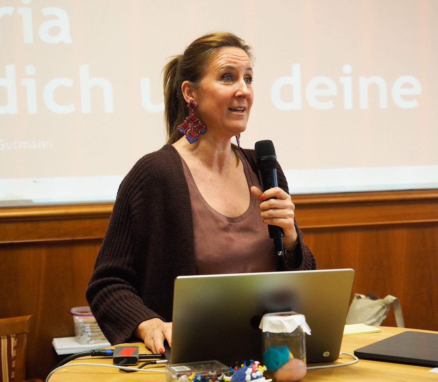 Eine Frau steht an einem Rednerpult, hält ein Mikrofon in der Hand und hält eine Rede auf dem Kreativwirtschaftsforum. Sie trägt ein braunes Oberteil und große Ohrringe und steht hinter einem Laptop in einem Klassenzimmer oder Hörsaal. Auf der Leinwand hinter ihr wird Text projiziert.