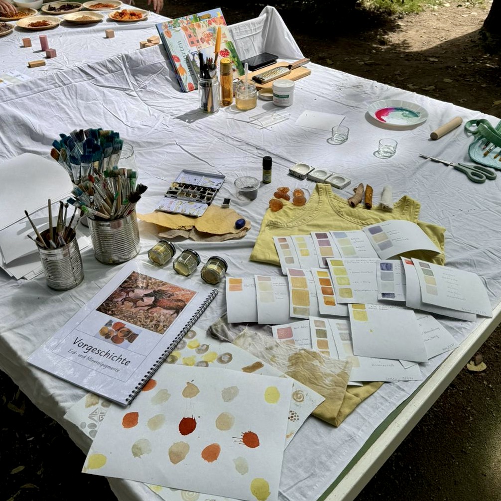 Art supplies, color swatches, brushes, notebooks, and glasses are spread out on a white tablecloth outdoors, suggesting an art workshop or a creative activity currently in progress—a true “community of creative worlds,” inspired by Yunuyei’s lively spirit.