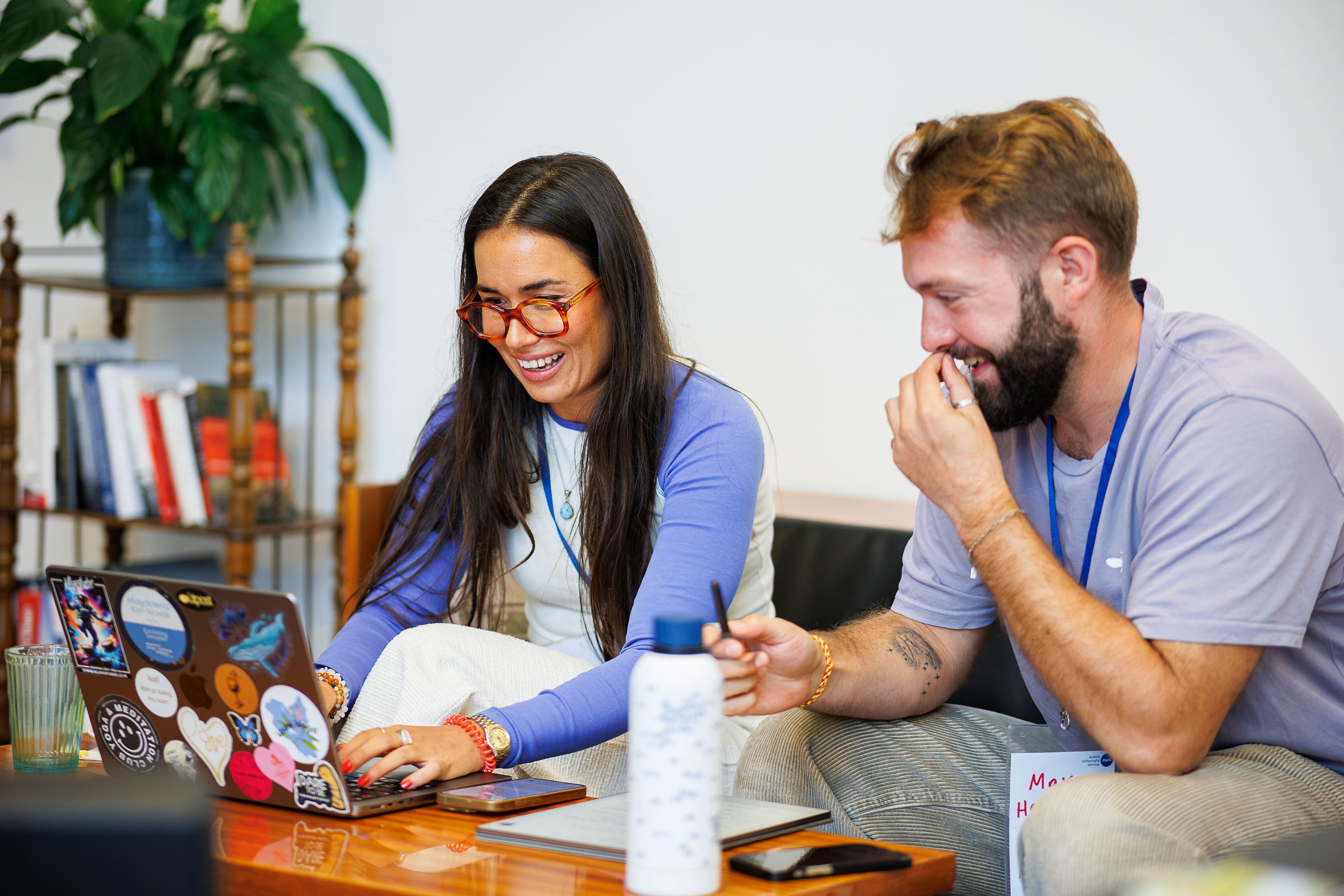 Eine Frau und ein Mann sitzen gemeinsam an einem Tisch, betrachten einen mit Aufklebern übersäten Laptop, lachen und teilen einen Moment persönlicher Erfahrung.