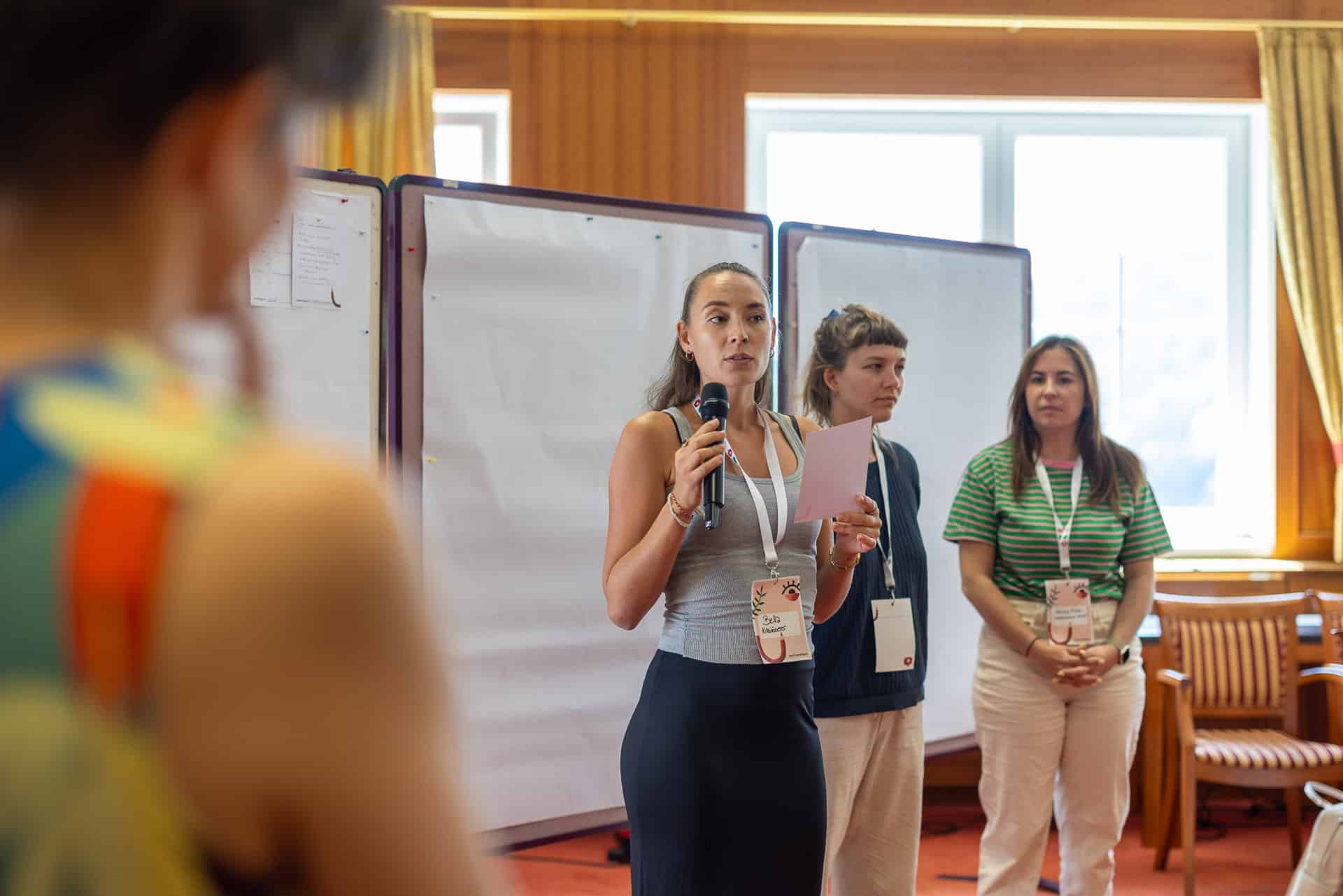 Eine Frau spricht während eines Workshops zur Logoentwicklung in ein Mikrofon, während sie ein Blatt Papier in der Hand hält; sie steht vor zwei Whiteboards, und zwei weitere Frauen hören ihr in der Nähe zu.
